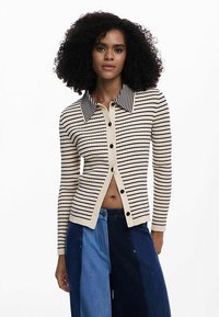 Cardigan cu dungi bej și bleumarin, cu guler ascuțit, textură răsucită și închidere frontală cu nasturi, purtat peste pantaloni din denim albastru cu tonuri diferite.