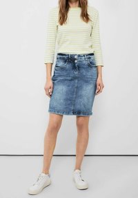 Falda de denim en azul medio con una silueta recta, dos bolsillos frontales y un cierre de botón visible. Combinada con una blusa de rayas verdes y zapatillas blancas.