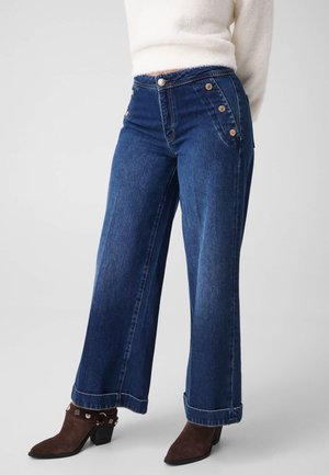 Femme portant un jean bleu taille haute à jambes larges avec des détails de boutons et des bottines marron à talons, associée à un pull blanc duveteux.