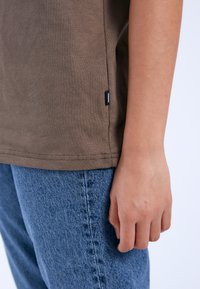 Camiseta de algodón marrón con un diseño ajustado, que presenta una etiqueta lateral y combinada con unos vaqueros de denim azul que muestran un acabado texturizado.