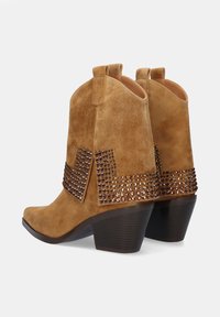 Botas de cowboy de ante marrón con un acento de chevrón cosido y remaches decorativos marrones en el talón, con una punta afilada y un tacón robusto.
