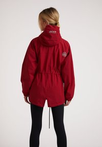 Rød vandtæt jakke med snoretræk i livet og hætte. Har elastiske manchetter og et logo-patch på den øverste ryg. Glat tekstur.