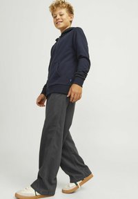 Mornarska zip majica s kapuco z sprednjimi žepi, v kombinaciji z ohlapnimi ogljeno sivimi hlačami in svetlimi supergami z zelenimi poudarki.