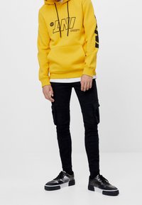 Żółta bluza z kapturem z czarnym graficznym napisem, czarne spodnie cargo z bocznymi kieszeniami i czarne niskie trampki ze srebrnymi akcentami.
