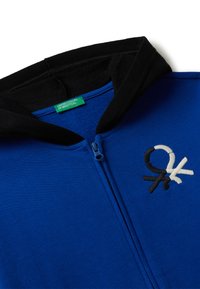 Sweat à zip bleu avec une capuche noire, logo brodé en blanc, noir et crème. Texture lisse, mélange de coton, avec une étiquette de marque verte.