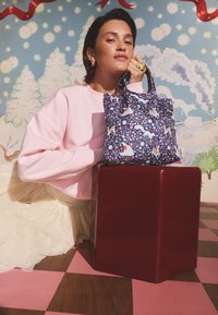 Maža tamsiai mėlyna rankinė su gėlių ir gyvūnų raštais. Turi du rankenas ir minkštą tekstūrą, pastatyta ant blizgančios raudonos kubo priešais žaismingą foną.