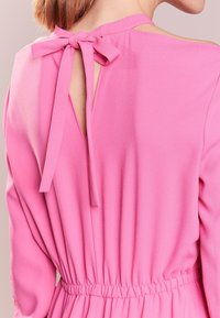 Rosa klänning med en knuten rosett på ryggen, med en öppen utskärning. Tillverkad i lätt tyg, har den en figurnära midja och långa ärmar.