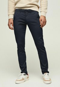 Marinblå slim-fit byxor i en bomullsblandning med en slät textur och standardfickor, i kombination med vita och beige sneakers.