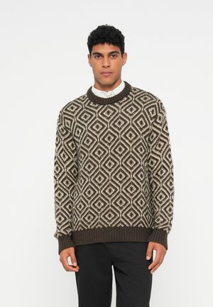Mann, der einen braun-beige gestreiften Strickpullover über einem weißen Hemd mit schwarzen Hosen trägt, steht vor einem schlichten weißen Hintergrund.