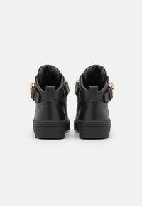 Ett par svarta höga sneakers med vadderade kragar och guldspännen, sedd bakifrån mot en vit bakgrund.