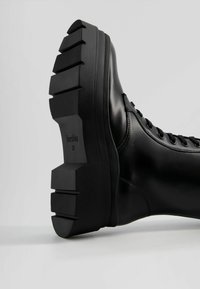 Botte en cuir noir avec une semelle en caoutchouc épaisse et rainurée. Dispose de lacets ronds et d'une texture lisse, étiquetée "Bershka 37" sur la semelle.