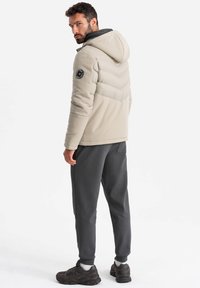 Giacca isolante beige con cappuccio, design a lavorazione a zig-zag e patch rotondo con logo sulla manica, abbinata a pantaloni grigio scuro e sneakers.