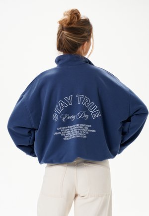 Femme aux cheveux en chignon portant un sweat-shirt bleu marine avec l'inscription « STAY TRUE Every Day » et un texte motivant dans le dos, associé à un pantalon clair.