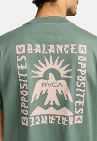 Green cotton t-shirt featuring a large graphic on the back with a sun, bird, and text "BALANCE OPPOSITES," in soft pink.
