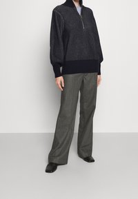 Person, die einen navy gestreiften Quarter-Zip-Pullover, graue weit geschnittene Hosen und schwarze eckige Schuhe trägt, steht vor einer schlichten Wand.