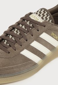 Καφέ σουέτ αθλητικό παπούτσι Adidas Spezial με λευκές ρίγες, υφασμένη γλώσσα, καφέ κορδόνια και ημιδιάφανη καουτσούκ σόλα.