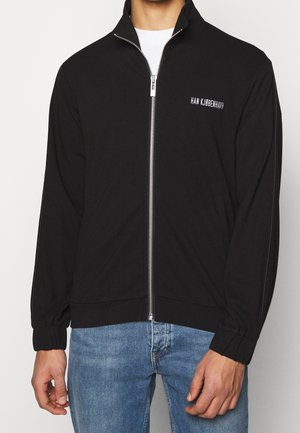 Hombre con chaqueta negra con cremallera y logo "HAN KJØBENHAVN" en el pecho y vaqueros azul claro sobre un fondo liso.
