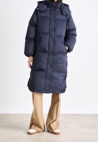 Marineblauer Steppmantel mit Kapuze, mit gesteppter Optik, Seitentaschen und Druckknopfverschluss. Kombiniert mit beigeweiten Hosen und Schuhen.