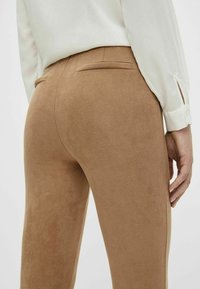 Brązowe elastyczne spodnie o miękkiej, teksturowanej powierzchni, z tylną kieszenią i dopasowanym krojem, zestawione z jasną bluzką.