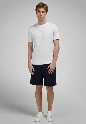 Young man standing wearing a white short-sleeve t-shirt, navy knee-length shorts, and white sneakers against a plain gray background.