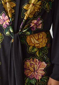 Tessuto nero con ricami floreali vivaci in rosa, giallo e verde. Il tessuto ha una texture morbida e un dettaglio annodato in vita.