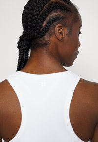 Balts sporta bez piedurknēm krekls ar apaļu kakla izgriezumu un sacīkšu muguru, ar nelielu logo augšējā mugurā. Tekstūras audums ar gludu apdari.