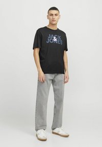 Mustvärviline t-särk, millel on sinine "JACK & JONES" logo. Kombineeritud hallide teksade ja valgete spordijalatsitega. Mudel seisab ühetaolise taustaga.