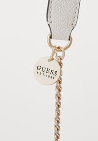 Bracelet en cuir gris clair avec un attachement en chaîne dorée. Présente une étiquette métallique ronde avec "GUESS EST. 1981" embossé en noir.