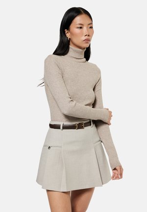 Kvinde med langt sort hår iført beige ribstrikket højhalset sweater og lysegråt plisseret mini-nederdel med mørkebrun bælte, der kigger sidelæns.