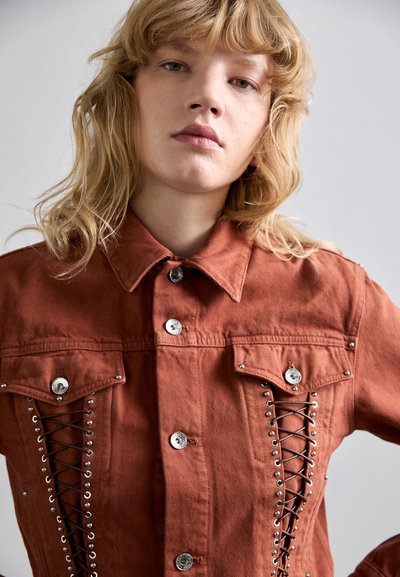 Jachetă din denim de culoare ruginie, cu butoni argintii, buzunare frontale și detalii cu șireturi și oțeluri pe laterale. Finisaj de material texturat.