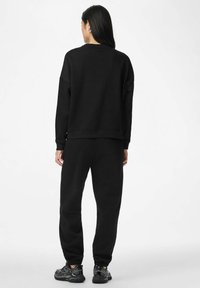 Moteris su ilgais tamsiais plaukais, dėvinti juodą džemperį ir sportines kelnes, stovinti atsukusi nugarą prieš paprastą baltą foną.