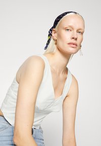 Top cropped branco com um design de decote em V. Modelo usa um lenço de padrão azul marinho e brincos de argola dourados com detalhes em pérola.