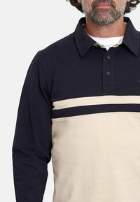 Polo de manga larga que presenta una parte superior en azul marino con una sección inferior en crema y dos rayas horizontales en azul marino en el pecho. Con cuello abotonado.