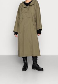 Parka oversized color caqui con capucha ajustable con cordón, bolsillos con cremallera y tejido texturizado. Combinada con botas de combate negras con cordones.