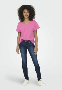 Lyserød V-hals kortærmet top parret med mørkeblå skinny jeans. Modellen står i hvide sneakers og viser et afslappet fit og blødt materiale.