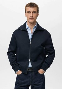 Giacca blu navy con cerniera, realizzata in tessuto in pile, con collo alto e polsini a coste, abbinata a una camicia azzurra e jeans scuri.