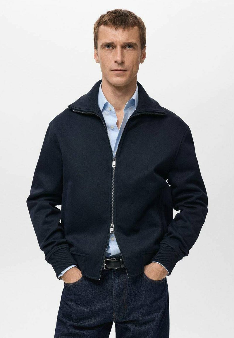 Giacca blu navy con cerniera, realizzata in tessuto in pile, con collo alto e polsini a coste, abbinata a una camicia azzurra e jeans scuri.