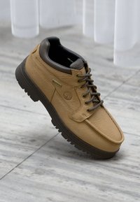 Kohvist tan värvi Gore-Tex matkajalats pruuni tald ja paelad, mis seisavad heledal hallil plaaditud põrandal pehmete varjudega.