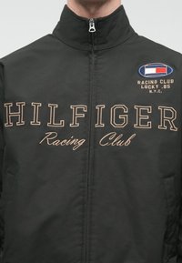 Jachetă neagră cu fermoar, având brodate în nuanțe de bej "HILFIGER" și "Racing Club". Include o etichetă cu logo în detalii albastre, albe și roșii.
