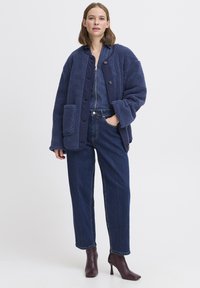 Veste en polaire bleu marine avec une fermeture à bouton, grandes poches et tissu texturé, associée à un pantalon en denim et des bottes à talons bordeaux.