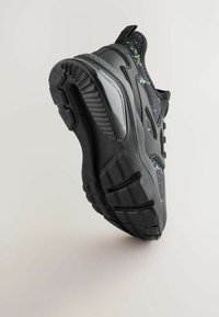 Sort sneakers med en struktureret gummisål, mesh-overdel med grønne sprøjte mønstre og forstærket hæl; skrå vinkel, der viser strukturen.