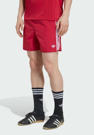 Person trägt rote Adidas-Shorts mit weißen Streifen, schwarze Adidas-Socken mit weißen Streifen und schwarze Adidas-Schuhe.