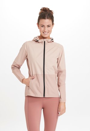 Ljusrosa zip-up jacka med huva, prydd med en svart dragkedja och elastiska muddarna, visad över en matchande outfit. Slätt, lätt material.