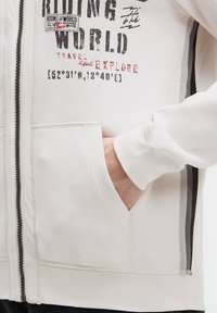 Weiße Zip-Hoodie mit bedruckten Grafiken, Fronttaschen und kontrastierenden grauen Streifen an den Seiten. Weiches Material, lockerer Schnitt.