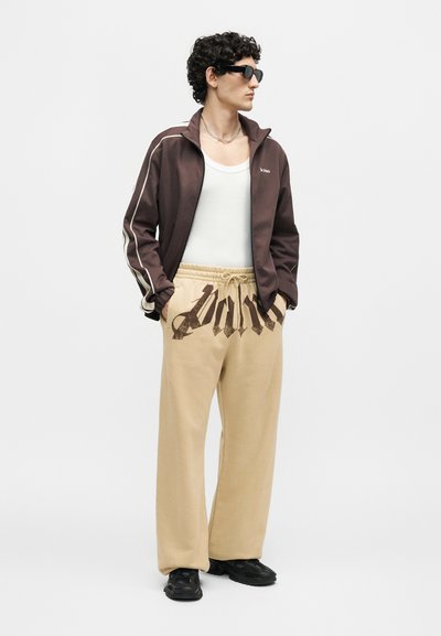Άνδρας που φοράει μαύρα γυαλιά ηλίου, σκούρο καφέ μπουφάν με φερμουάρ, λευκό τοπ, μπεζ παντελόνι με μεγάλα μαύρα γράμματα και μαύρα αθλητικά παπούτσια, στέκεται με τα χέρια στις τσέπες.