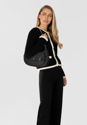 Borsa a mano in pelle nera dalla forma curva e texture liscia, con dettaglio fibbia, indossata sulla spalla con un maglione bianco e nero.