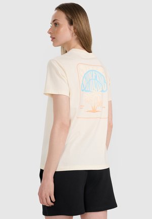 Kobieta o prostych jasnobrązowych włosach, ubrana w kremowy T-shirt z kolorowym nadrukiem o tematyce surfingu na plecach i czarne szorty, zwrócona w lewo.