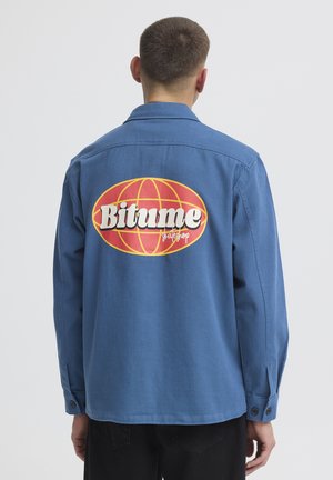 Personne portant une chemise bleue à boutons avec un grand logo globe rouge et jaune et le texte "Bitume skateshop" dans le dos.