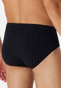 Homme portant un slip noir avec un motif de grille bleu clair subtil, montré du bas du dos jusqu'au milieu de la cuisse sur un fond uni.