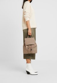 Beige läderryggsäck med klaffstängning, två dragkedjefickor och en strukturerad form. Detaljer inkluderar kontrasterande sömmar och ett handtag i toppen.